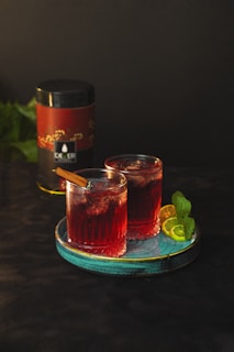 Two glasses of a red cocktail are placed on a turquoise plate. Each glass is garnished with a cinnamon stick. Behind the glasses, there is a cylindrical container with a red and black label. Lime slices and mint leaves decorate the plate.