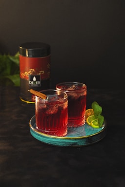 Two glasses of a red cocktail are placed on a turquoise plate. Each glass is garnished with a cinnamon stick. Behind the glasses, there is a cylindrical container with a red and black label. Lime slices and mint leaves decorate the plate.