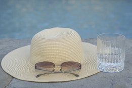 A straw hat with a wide brim is placed on a stone surface next to a pair of sunglasses and an empty glass. The background features a blurred view of a swimming pool, creating a relaxed, summertime atmosphere.