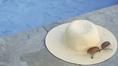 Classic wayfarer sunglasses perched on a sunhat beside a pool.