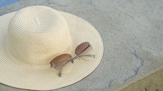 A wide-brimmed straw hat is placed on a stone surface with a pair of sunglasses resting on top. The scene conveys a sense of leisure, possibly suggesting a day out in the sun.