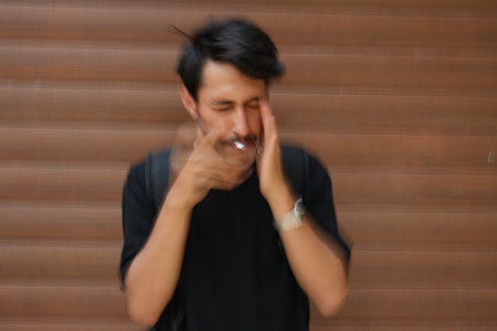 A person is standing in front of a wooden shutter, with motion blur making the face and hands appear out of focus. They are wearing a black shirt and a watch, with their eyes closed and hands touching their face. A cigarette is held between their fingers.