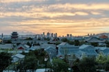 A scenic view of traditional Chinese architecture among lush greenery, set against a backdrop of a modern city skyline at sunset. The sky is filled with warm hues of orange and yellow clouds, creating a striking contrast with the grey-blue rooftops.
