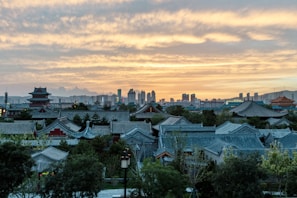 A scenic view of traditional Chinese architecture among lush greenery, set against a backdrop of a modern city skyline at sunset. The sky is filled with warm hues of orange and yellow clouds, creating a striking contrast with the grey-blue rooftops.