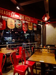 A cozy pizzeria interior with red chairs and wooden tables. Above, a bright red sign promotes reasons to love pizza. The lighting is warm, with decorative pendant lights hanging from the ceiling. Outside, through the glass, a street view with parked cars and neon signs is visible.