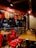 A cozy pizzeria interior with red chairs and wooden tables. Above, a bright red sign promotes reasons to love pizza. The lighting is warm, with decorative pendant lights hanging from the ceiling. Outside, through the glass, a street view with parked cars and neon signs is visible.