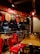A cozy pizzeria interior with red chairs and wooden tables. Above, a bright red sign promotes reasons to love pizza. The lighting is warm, with decorative pendant lights hanging from the ceiling. Outside, through the glass, a street view with parked cars and neon signs is visible.
