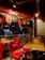 A cozy pizzeria interior with red chairs and wooden tables. Above, a bright red sign promotes reasons to love pizza. The lighting is warm, with decorative pendant lights hanging from the ceiling. Outside, through the glass, a street view with parked cars and neon signs is visible.