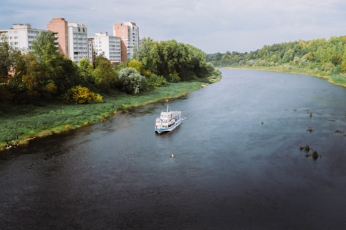 A river meanders through a landscape with lush green trees lining its banks. On one side, residential buildings with a utilitarian design rise above the treetops. A small boat sails along the river, creating gentle ripples in the water.