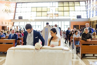 A bride and groom are standing at the altar of a church, engaged in a marriage ceremony. The bride is wearing a white gown and veil, holding a small bouquet of flowers. The groom is dressed in a formal suit. The church is filled with seated guests observing the ceremony, with large windows and chandeliers providing an elegant setting.