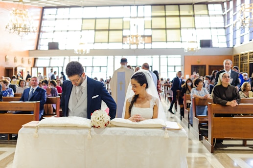 A bride and groom are standing at the altar of a church, engaged in a marriage ceremony. The bride is wearing a white gown and veil, holding a small bouquet of flowers. The groom is dressed in a formal suit. The church is filled with seated guests observing the ceremony, with large windows and chandeliers providing an elegant setting.