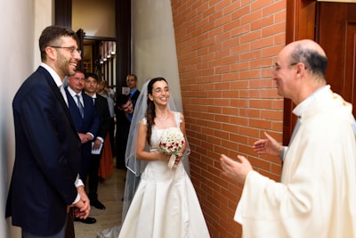 A bride in a white wedding dress is standing next to a groom in a dark suit, both smiling, with the bride holding a bouquet of red and white flowers. A priest is standing across from them, engaging in conversation. Several guests in formal attire are visible in the background, standing in a hallway with a brick wall.