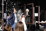 A newlywed couple, dressed in a suit and a wedding dress, is exiting a building surrounded by people. They are showered with what appears to be flower petals or confetti. The bride holds a bouquet of white and red flowers. Several people are celebrating around them, and the scene is lively and joyful.