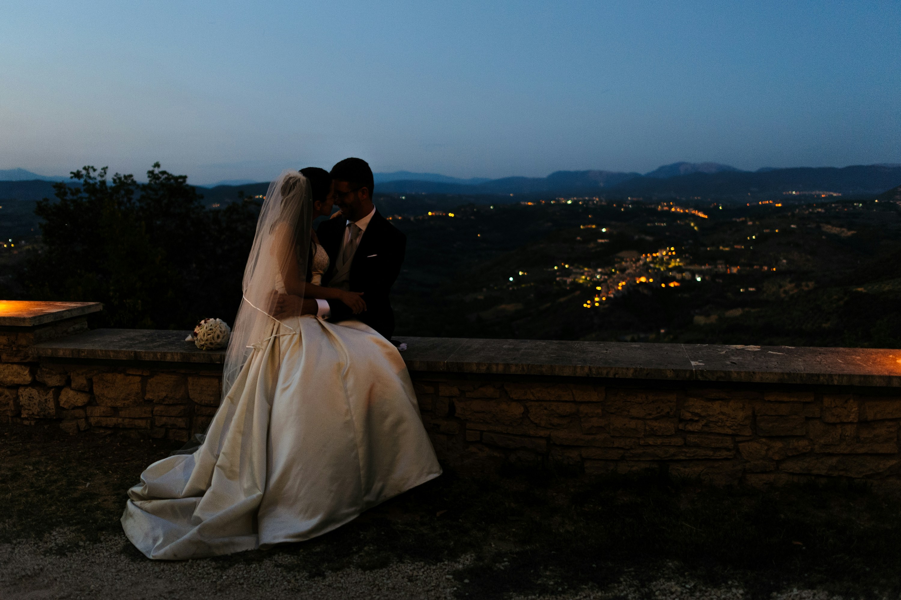 a bride and groom sitting on a stone wall at night