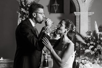 A couple is captured in a toast, sharing a sip of champagne while holding each other's arms. They are elegantly dressed, with the man in a suit and the woman in a bridal gown adorned with flowers. The background features arches and floral arrangements, suggesting a wedding setting, along with candles adding to the ambiance.