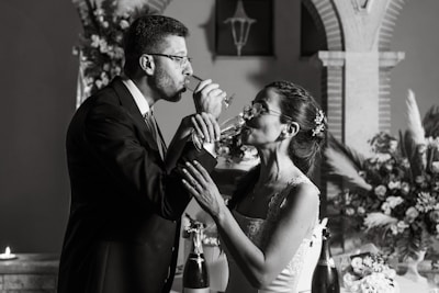 A couple is captured in a toast, sharing a sip of champagne while holding each other's arms. They are elegantly dressed, with the man in a suit and the woman in a bridal gown adorned with flowers. The background features arches and floral arrangements, suggesting a wedding setting, along with candles adding to the ambiance.