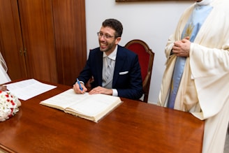 A person in a formal suit is seated at a wooden desk, smiling broadly while signing a document. Their pen is poised above an open book, which is likely a registry or official ledger. Next to the seated person, partially visible, someone wearing a light-colored robe stands, possibly observing the signing process. In the foreground, the table is adorned with a bouquet of white and red flowers, and there are papers laid out beside the book.