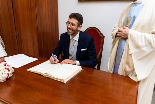 A person in a formal suit is seated at a wooden desk, smiling broadly while signing a document. Their pen is poised above an open book, which is likely a registry or official ledger. Next to the seated person, partially visible, someone wearing a light-colored robe stands, possibly observing the signing process. In the foreground, the table is adorned with a bouquet of white and red flowers, and there are papers laid out beside the book.