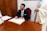 A person in a formal suit is seated at a wooden desk, smiling broadly while signing a document. Their pen is poised above an open book, which is likely a registry or official ledger. Next to the seated person, partially visible, someone wearing a light-colored robe stands, possibly observing the signing process. In the foreground, the table is adorned with a bouquet of white and red flowers, and there are papers laid out beside the book.