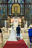A couple is standing in a church in front of an altar, adorned with a floral arrangement. The church features ornate stained glass windows and intricate architectural details. A priest or officiant stands at the side, conducting the ceremony, while guests are seated nearby.