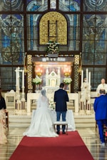 A couple is standing in a church in front of an altar, adorned with a floral arrangement. The church features ornate stained glass windows and intricate architectural details. A priest or officiant stands at the side, conducting the ceremony, while guests are seated nearby.