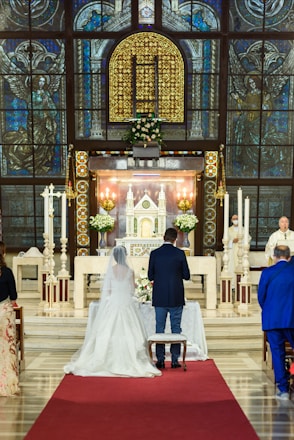 A couple is standing in a church in front of an altar, adorned with a floral arrangement. The church features ornate stained glass windows and intricate architectural details. A priest or officiant stands at the side, conducting the ceremony, while guests are seated nearby.