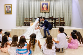 A bride and groom share a kiss while seated on a small stage in front of a group of children. The bride is in a white gown and the groom is in a dark suit. A portrait and another painting hang on the wall behind them, with a table and chairs beneath the artwork. The children, dressed in various colorful outfits, sit on the floor facing the couple.