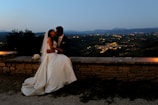 A couple in wedding attire sits on a stone wall, sharing a kiss against a backdrop of a scenic landscape at dusk. The bride is wearing a white gown with a veil, and the groom is in a suit. The distant view includes a softly lit valley with twinkling lights, surrounded by hills.