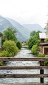A picturesque scene featuring a small river flowing gently through a village, bordered by lush green trees and rustic houses. In the background, majestic mountains rise, partly obscured by light clouds. The scene is viewed from a wooden bridge, adding a quaint charm to the setting.