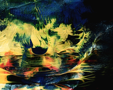A textured and abstract composition featuring swirling patterns of green, blue, yellow, and red. The colors blend together, creating shapes that resemble nature-inspired forms or surreal landscapes. The paint appears layered and dynamic, with a glossy finish reflecting light.