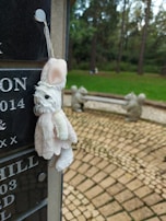 A plush toy resembling a small animal, possibly a rabbit, hangs from a string attached to a dark granite headstone with engraved letters and dates. In the background, there is a park-like setting with green grass, large trees, and a stone bench flanked by sculptures of squirrels.
