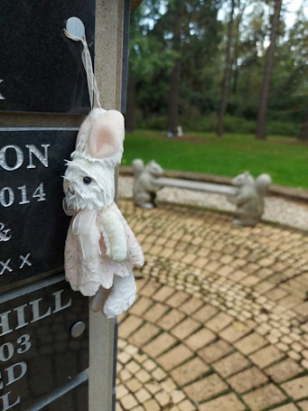 A plush toy resembling a small animal, possibly a rabbit, hangs from a string attached to a dark granite headstone with engraved letters and dates. In the background, there is a park-like setting with green grass, large trees, and a stone bench flanked by sculptures of squirrels.