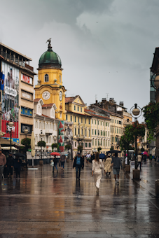 A rainy day scene in a European city with a prominent clock tower. People are walking with umbrellas along a wet street, and the architecture features colorful historic buildings. The overcast sky adds to the moody atmosphere.