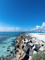 A scenic coastal view featuring a long row of large, square concrete blocks forming a barrier along the shoreline. The blocks extend towards the horizon, with clear, turquoise water on one side and a white sandy beach on the other. The sky is bright blue with scattered fluffy clouds.