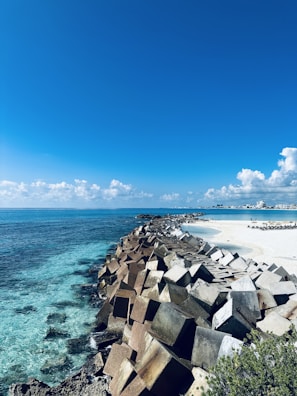 A scenic coastal view featuring a long row of large, square concrete blocks forming a barrier along the shoreline. The blocks extend towards the horizon, with clear, turquoise water on one side and a white sandy beach on the other. The sky is bright blue with scattered fluffy clouds.