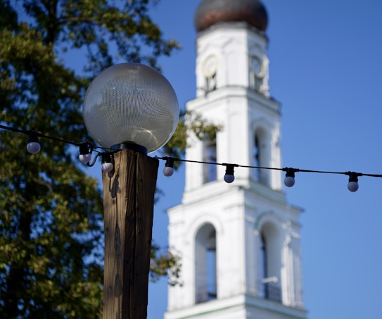 A wooden post supports a spherical street lamp, with strings of round bulbs attached. In the background, a white bell tower with arched windows and a dark dome stands tall. Green foliage is visible on the trees, set against a clear blue sky.