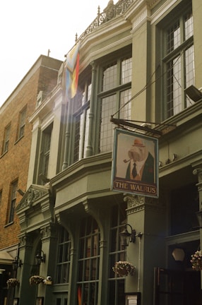 An ornate building features large, arched windows and a traditional pub sign that reads 'The Walrus.' A multicolored pride flag hangs near the top corner. The facade is detailed with columns and decorative molding, and potted flowers hang by the entrance.