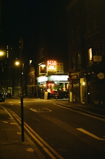 A dimly lit urban street scene at night featuring a pub with a bright neon sign. The street is mostly empty, with a few parked cars and streetlights casting a warm glow. The buildings are tall and closely packed, typical of a city environment.