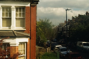 A residential street featuring red brick buildings with white window frames, parked cars lining the road, and trees with lush green foliage. The sky is partly cloudy, and there is a tall street lamp visible. The scene presents a typical urban neighborhood setting.