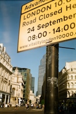 A street scene in London. A large yellow sign provides information about a road closure for an event. The background includes historic and modern buildings and a few people walking on the street. The sky is clear and blue.