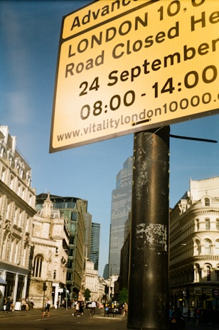 A street scene in London. A large yellow sign provides information about a road closure for an event. The background includes historic and modern buildings and a few people walking on the street. The sky is clear and blue.