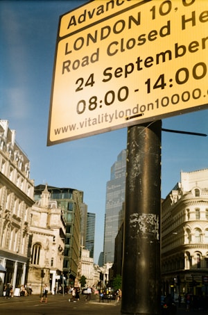 A street scene in London. A large yellow sign provides information about a road closure for an event. The background includes historic and modern buildings and a few people walking on the street. The sky is clear and blue.