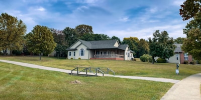 A suburban scene with a single-story house surrounded by well-maintained grass and trees. The house is painted in a light color with a dark roof, featuring a small porch with columns. A concrete pathway and a metal railing lead towards the house. The sky is partly cloudy with patches of blue peeking through.