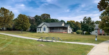 A suburban scene with a single-story house surrounded by well-maintained grass and trees. The house is painted in a light color with a dark roof, featuring a small porch with columns. A concrete pathway and a metal railing lead towards the house. The sky is partly cloudy with patches of blue peeking through.