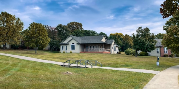 A suburban scene with a single-story house surrounded by well-maintained grass and trees. The house is painted in a light color with a dark roof, featuring a small porch with columns. A concrete pathway and a metal railing lead towards the house. The sky is partly cloudy with patches of blue peeking through.