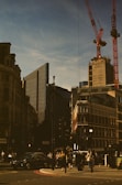 Urban scene featuring a city intersection with modern and historic buildings. Construction cranes are visible above the skyline, and various pedestrians can be seen walking on the street. Vehicles are waiting at the traffic lights under a clear blue sky.