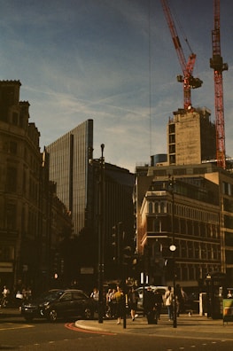 Urban scene featuring a city intersection with modern and historic buildings. Construction cranes are visible above the skyline, and various pedestrians can be seen walking on the street. Vehicles are waiting at the traffic lights under a clear blue sky.