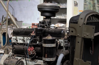 The image depicts a vintage engine in a workshop setting, with visible components such as wires, tubes, and metal parts. The surrounding environment includes shelving and diagrams on the walls, suggesting a repair or restoration area.