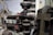 The image depicts a vintage engine in a workshop setting, with visible components such as wires, tubes, and metal parts. The surrounding environment includes shelving and diagrams on the walls, suggesting a repair or restoration area.