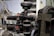 The image depicts a vintage engine in a workshop setting, with visible components such as wires, tubes, and metal parts. The surrounding environment includes shelving and diagrams on the walls, suggesting a repair or restoration area.